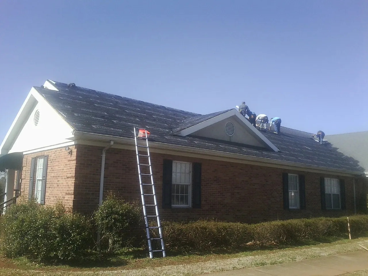 Crew tearing shingles off a brick ranch roof with a ladder set — Emergency Roof Repair work by our crew in Ravenel