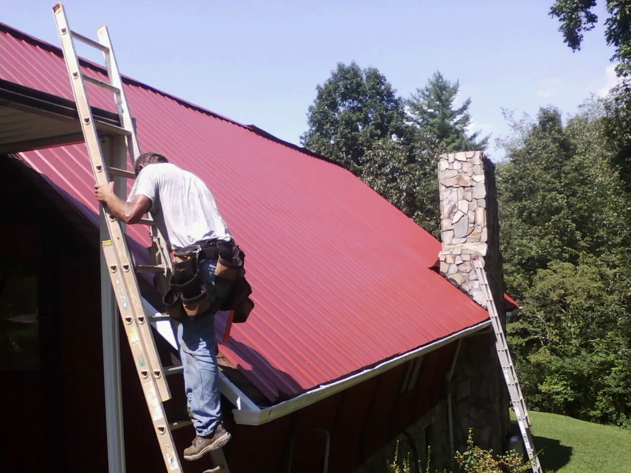 Roofer on a ladder beside a red metal roof and stone chimney — Soffit & Fascia Repair by Roofing Brothers in Ravenel